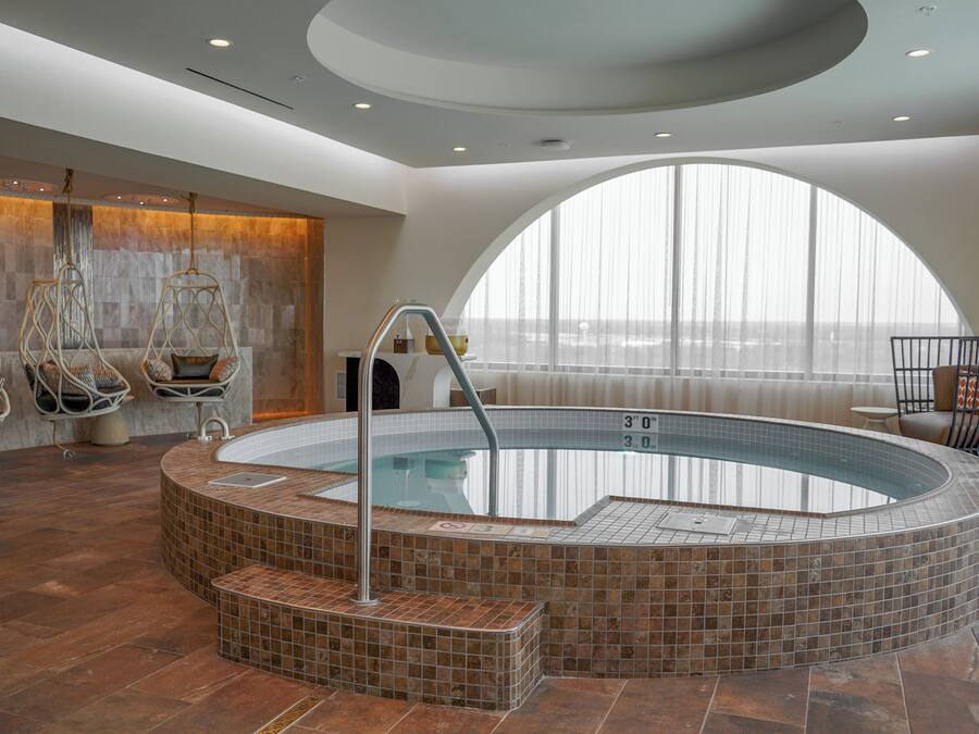 A modern indoor swimming pool with a tiled edge, a seating area with chairs, and a large window providing a view.