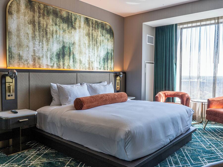 Une chambre d’hôtel avec un grand lit, une tête de lit agrémentée d’un décor mural texturé, une table de chevet, un bureau, deux chaises et une grande fenêtre avec des rideaux.