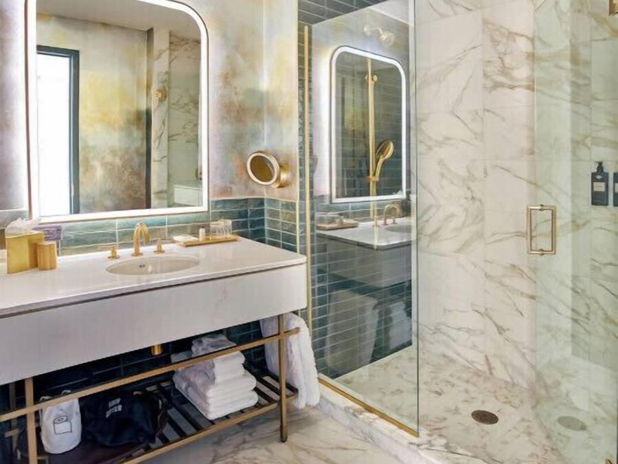 Een badkamer met een marmeren vloer, een witte wastafel met gouden details, een grote spiegel en een douchecabine met glazen wanden.