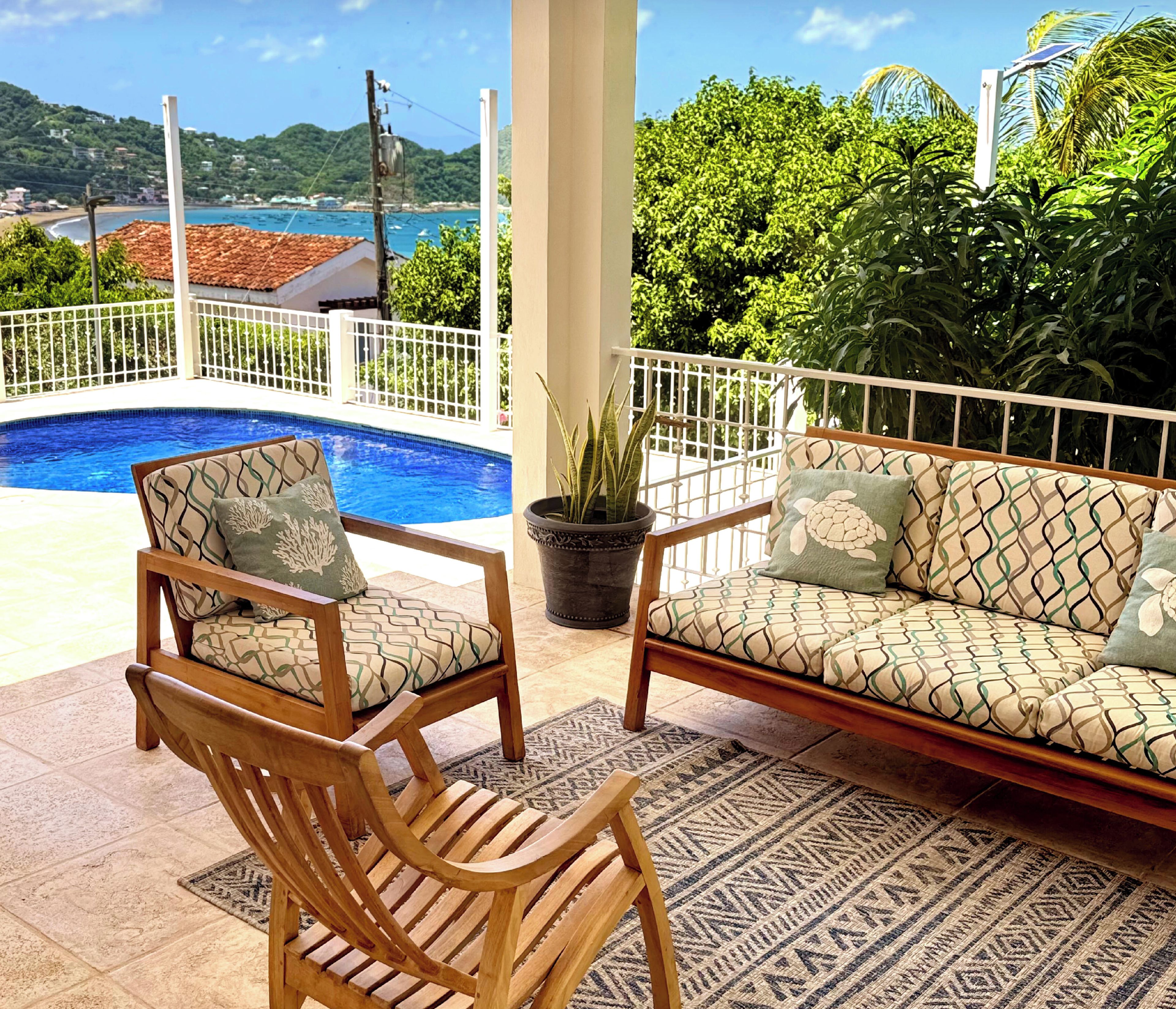 A balcony with a pool, wooden furniture, and a view of the sea and hills.