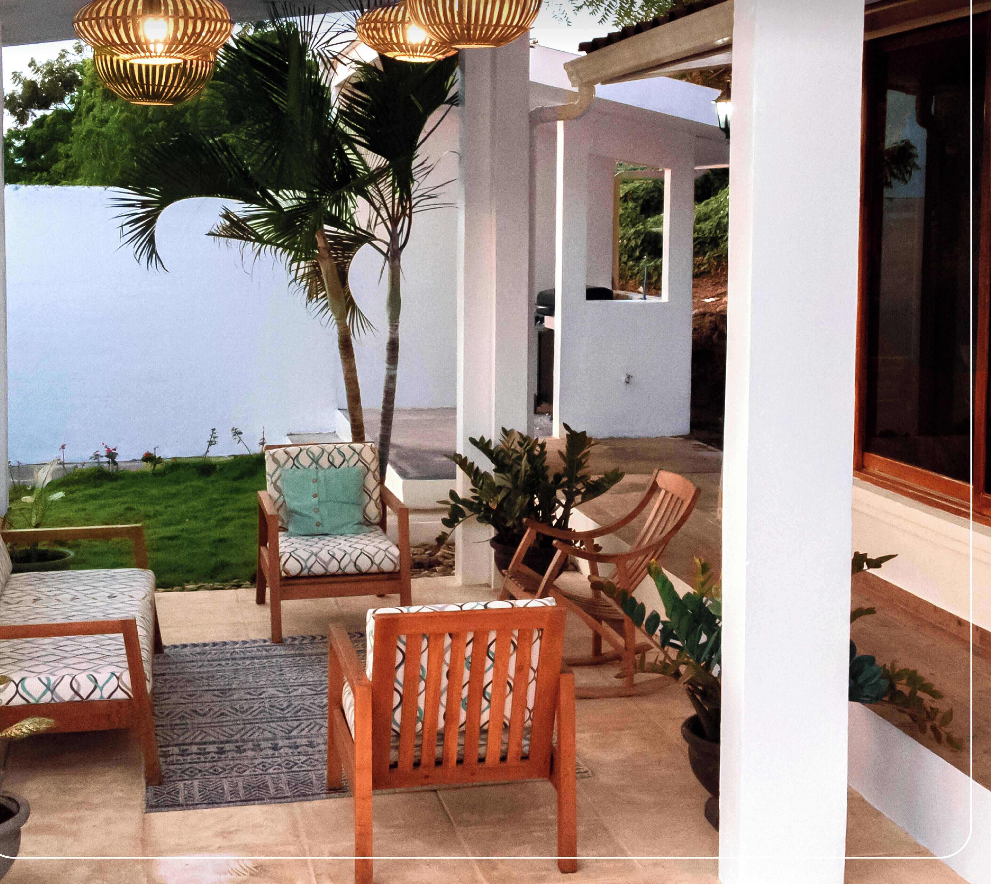 An outdoor patio with wooden furniture, a palm tree, and a view of the sea.