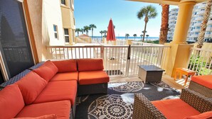 A balcony with a sofa and wicker furniture, overlooking a beach and palm trees. - Atlantic Villas 103 (New Smyrna Beach)