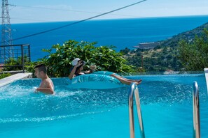 En swimmingpool med krystalklart, blåt vand, en person i en blå, oppustelig ring, og hav og bakker i baggrunden.