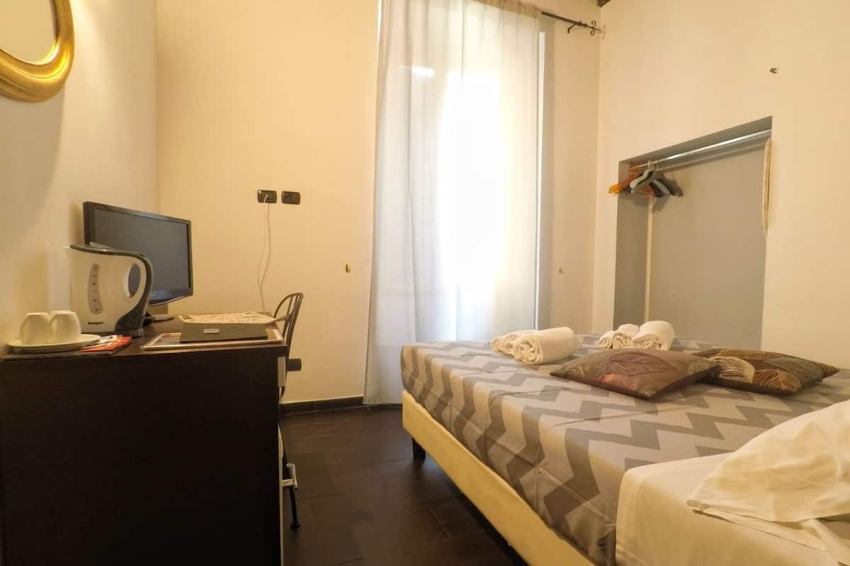 Quarto de hotel com cama, escrivaninha com computador e chaleira, janela com cortinas e espelho na parede.