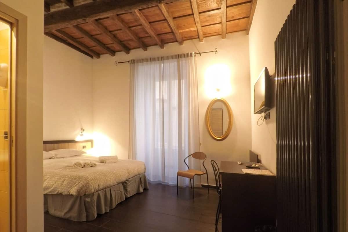 Quarto de hotel com cama, escrivaninha, cadeira, televisão e espelho.