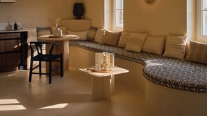 Ett mysigt vardagsrum med en böjd soffa, ett litet runt bord och en stol. Det finns ett fönster som släpper in naturligt ljus och en vägglampa.