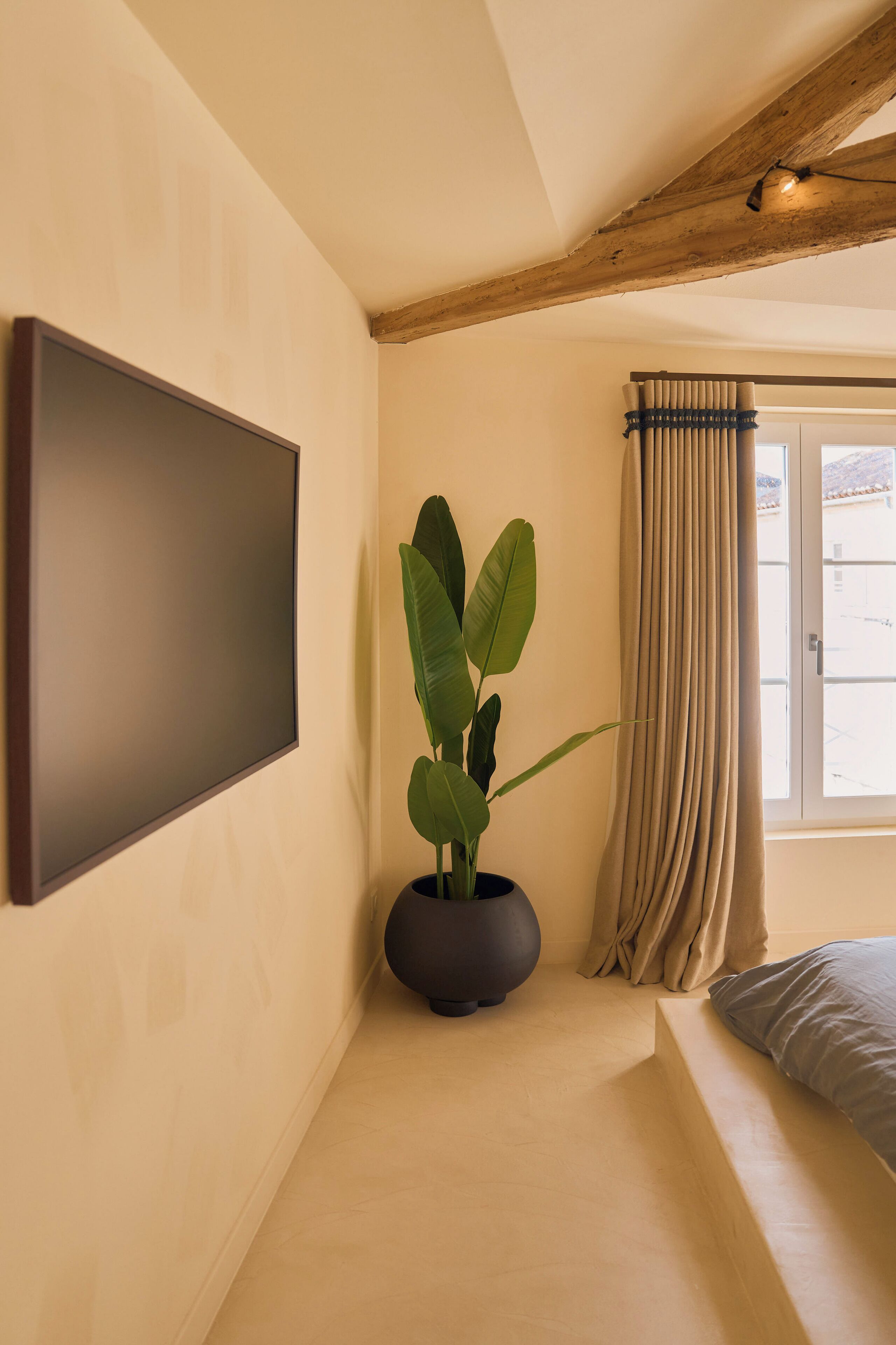 Une pièce dotée d’un grand téléviseur à écran plat fixé au mur, d’une plante en pot, d’une fenêtre avec des rideaux et d’un lit avec un oreiller.