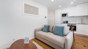 A modern apartment with a beige sofa, teal cushions, a wooden floor, a small round table, and a kitchen area with white cabinets and appliances. - GuestReady - Restful Getaway in Matosinhos (Matosinhos)