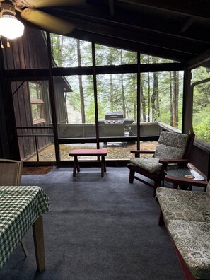 A covered outdoor seating area with a table and chairs, a view of a forest through large windows, and a barbecue grill outside. - Roomy, Rustic Cabin-4BR, Modern Amenities-Rothrock St Forest near State College (Petersburg)