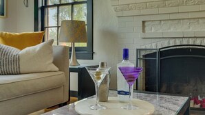 A living room with a sofa, a round table with a floral tablecloth, a martini glass, a bottle of vodka, and a lamp. - NEW Old Southwest Gem, Patio, Grill, Smart TV, 2 Desks + Fitness Equipment

 (Reno)