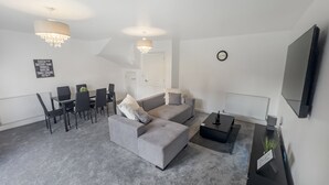 A modern living room with a grey sofa, a dining table with chairs, a coffee table, a TV mounted on the wall, and a chandelier.
