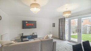A modern living room with a large flat-screen TV, a sofa, a coffee table, and a dining area with chairs.