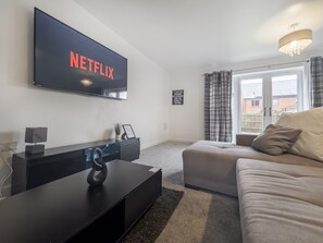 A modern living room with a large flat-screen TV mounted on the wall, a dark entertainment unit, a beige sofa, and a window with curtains. - Sleeps 8 With Garden and Pet Friendly (Ashford)