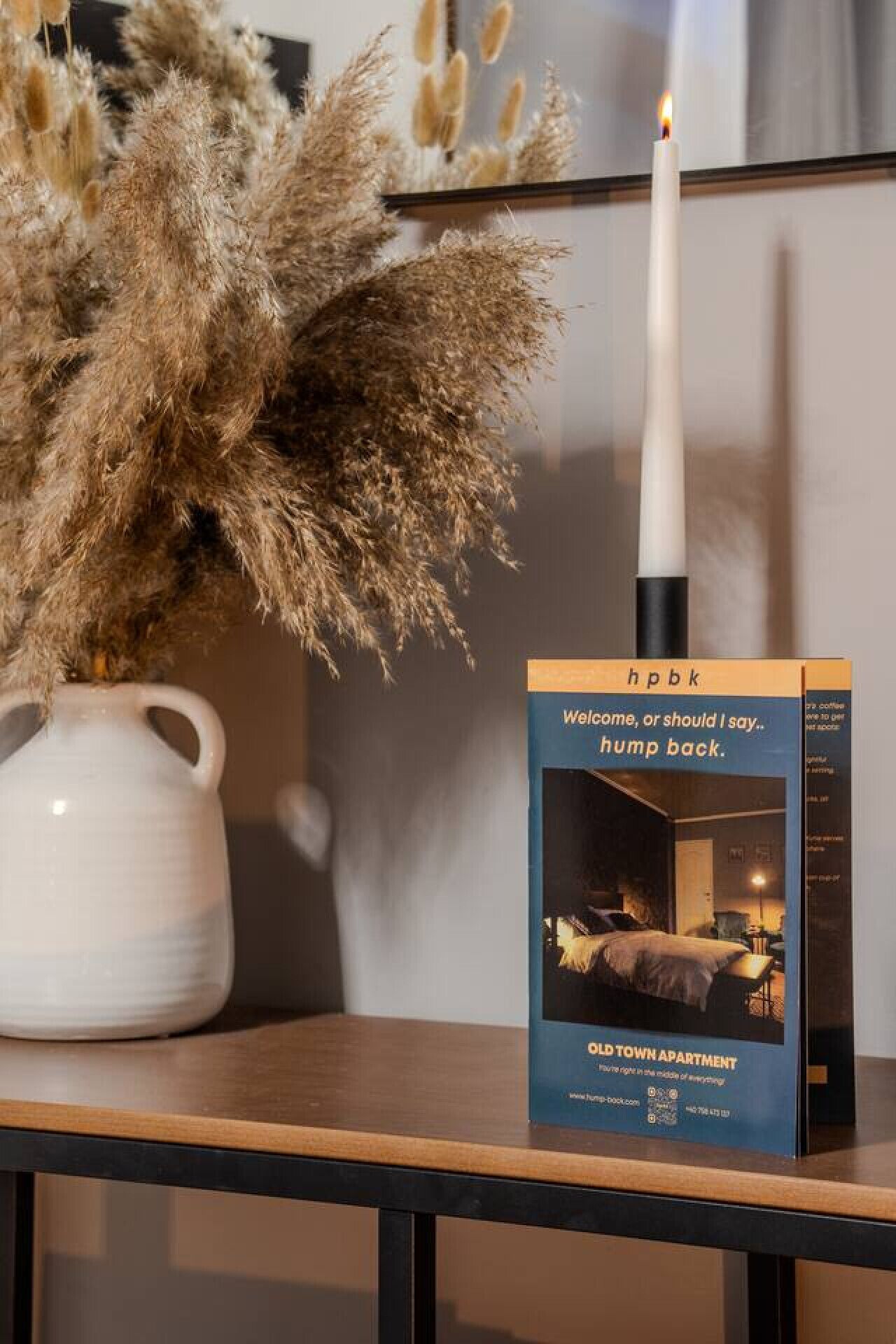 A candle, pampas grass, and a book titled 'hpbk' on a table.