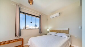 A bedroom with a bed, a bench, a window with curtains, an air conditioning unit, and a city view.