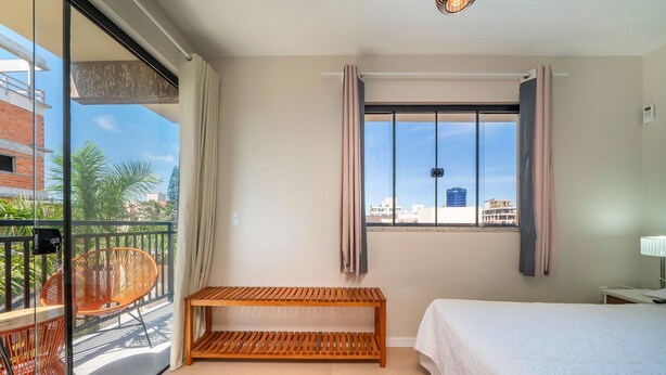 A hotel room with a bed, a wooden bench, a balcony with wicker chairs, and a view of the city.
