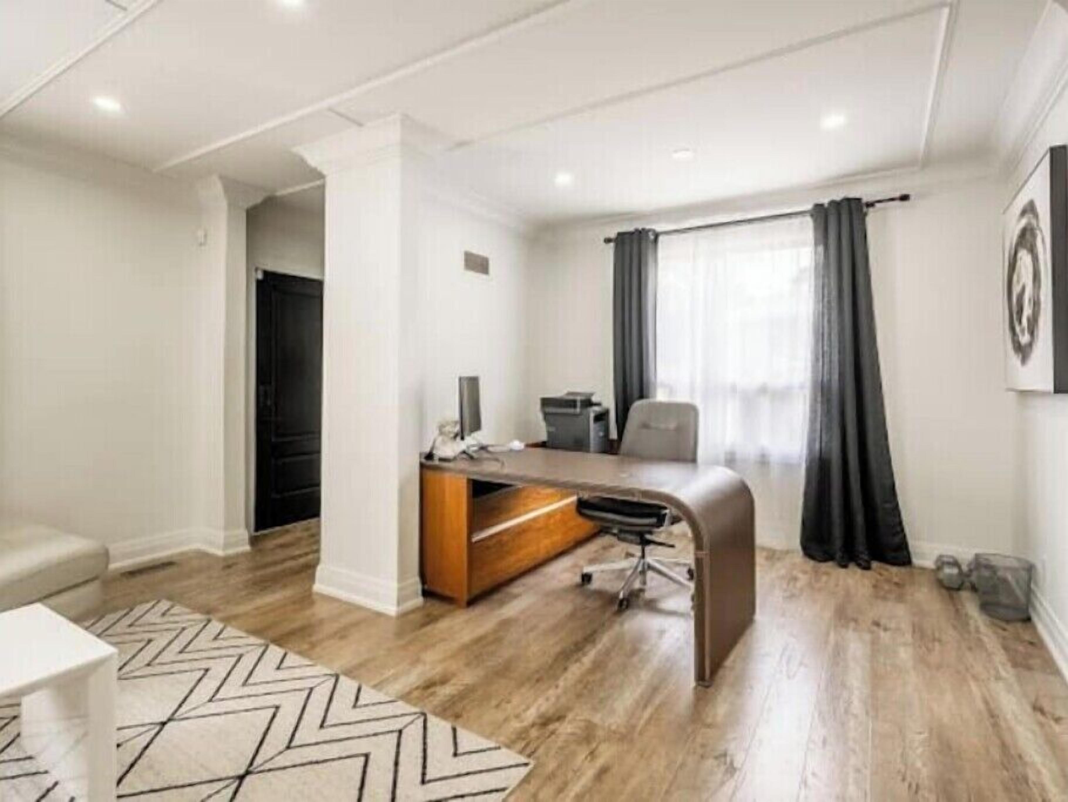 Un espacio de oficina moderno con escritorio, silla y ordenador. Hay una ventana con cortinas, una alfombra estampada y una puerta que da a otra habitación.