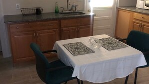 Een kleine keuken met houten kastjes, een spoelbak en een raam. Er staat een tafel met een witte tafelkleed en twee stoelen.