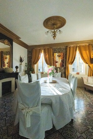 Ein Esszimmer mit einem runden Tisch, weißen Stühlen und einer Chandelieber. Es gibt einen Kamin, ein Sofa und ein Fenster mit Vorhängen.