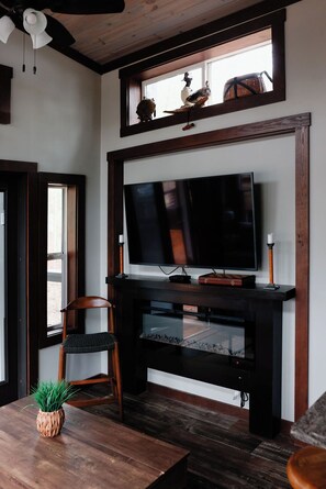 Una sala moderna con un televisor de pantalla plana grande montado en un mueble de entretenimiento de madera oscura, una silla de madera, una mesita con una planta y una ventana decorada con adornos en el alféizar.