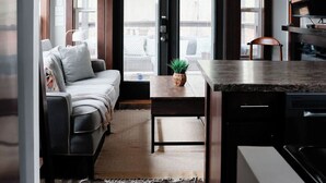 A modern kitchen with a wooden ceiling and a ceiling fan, a dining area with a table and chairs, and a living room with a sofa and a plant.