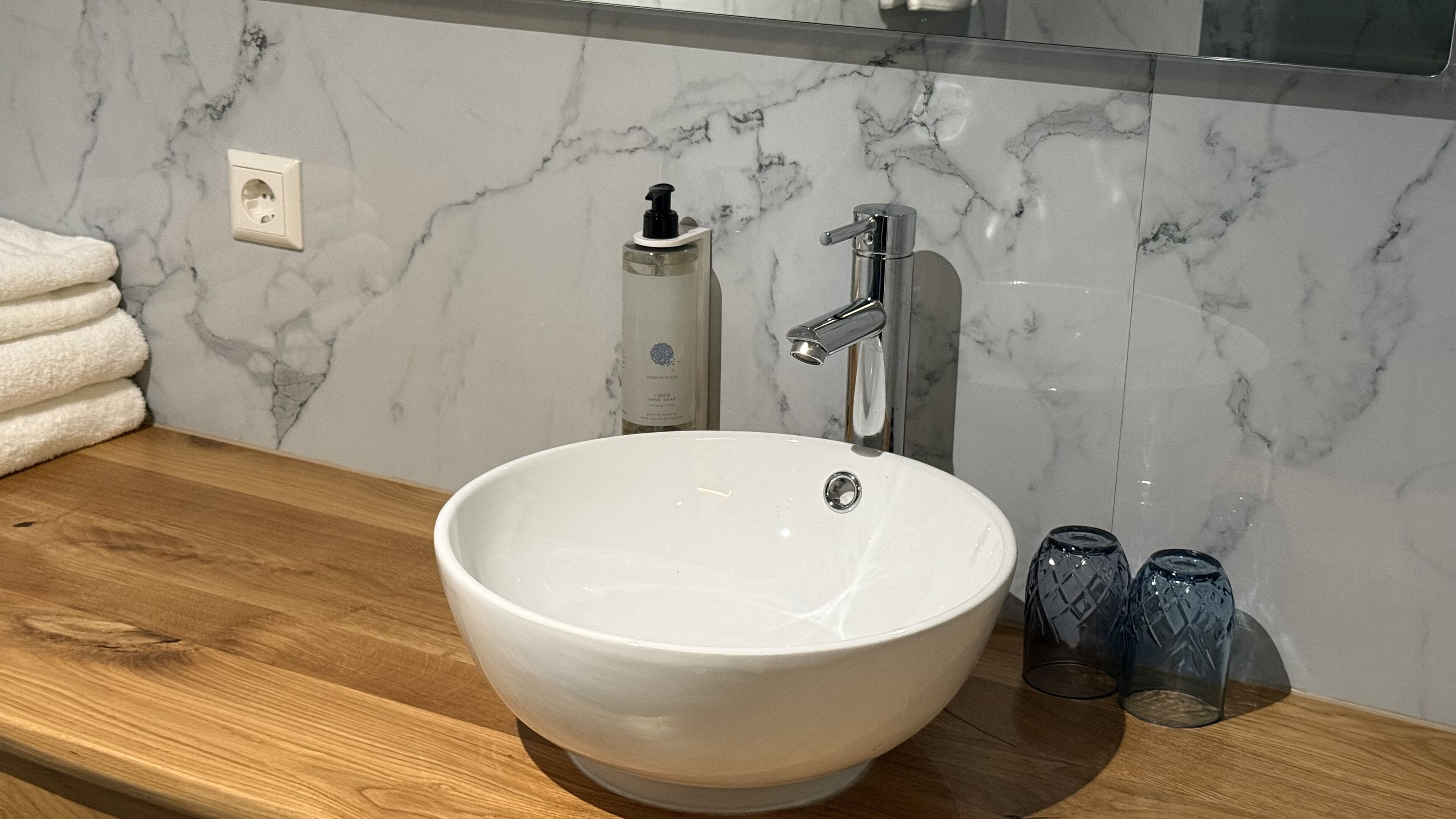 Un baño con lavamanos de mármol blanco, encimera de madera, un espejo y dos botellas de vidrio.
