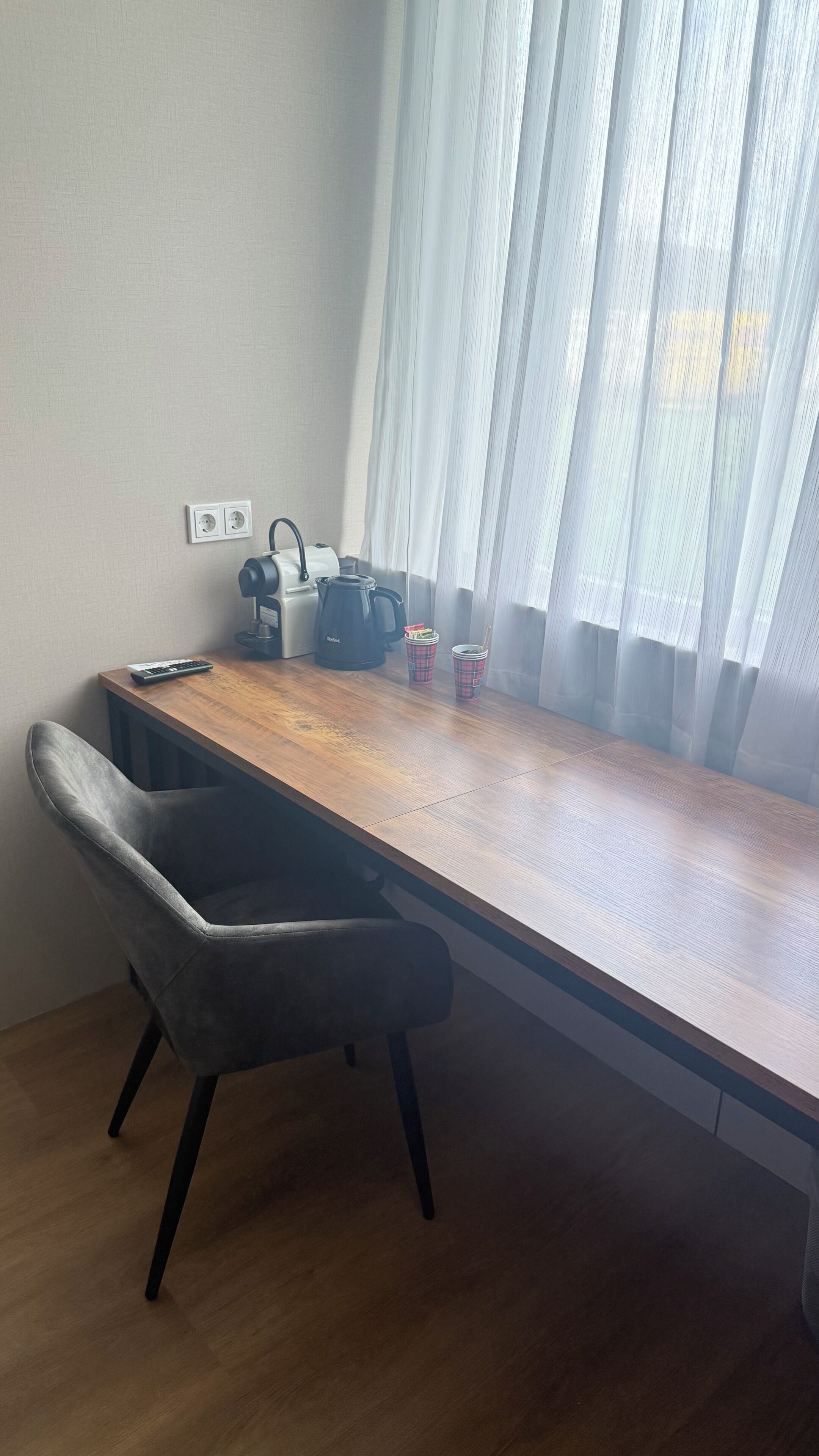 A room with a wooden desk, a black coffee maker, two plaid cups, a remote control, and a grey chair.