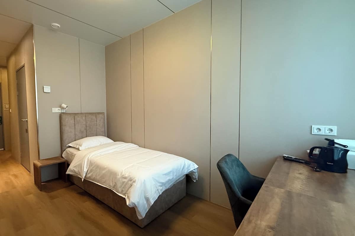 A small, neatly arranged bedroom with a bed, a chair, a desk, and a coffee maker.