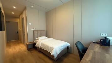 A small, neatly arranged bedroom with a bed, a chair, a desk, and a coffee maker.