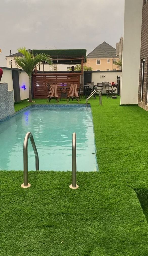 A swimming pool with a grassy deck, a covered seating area, and a building in the background.