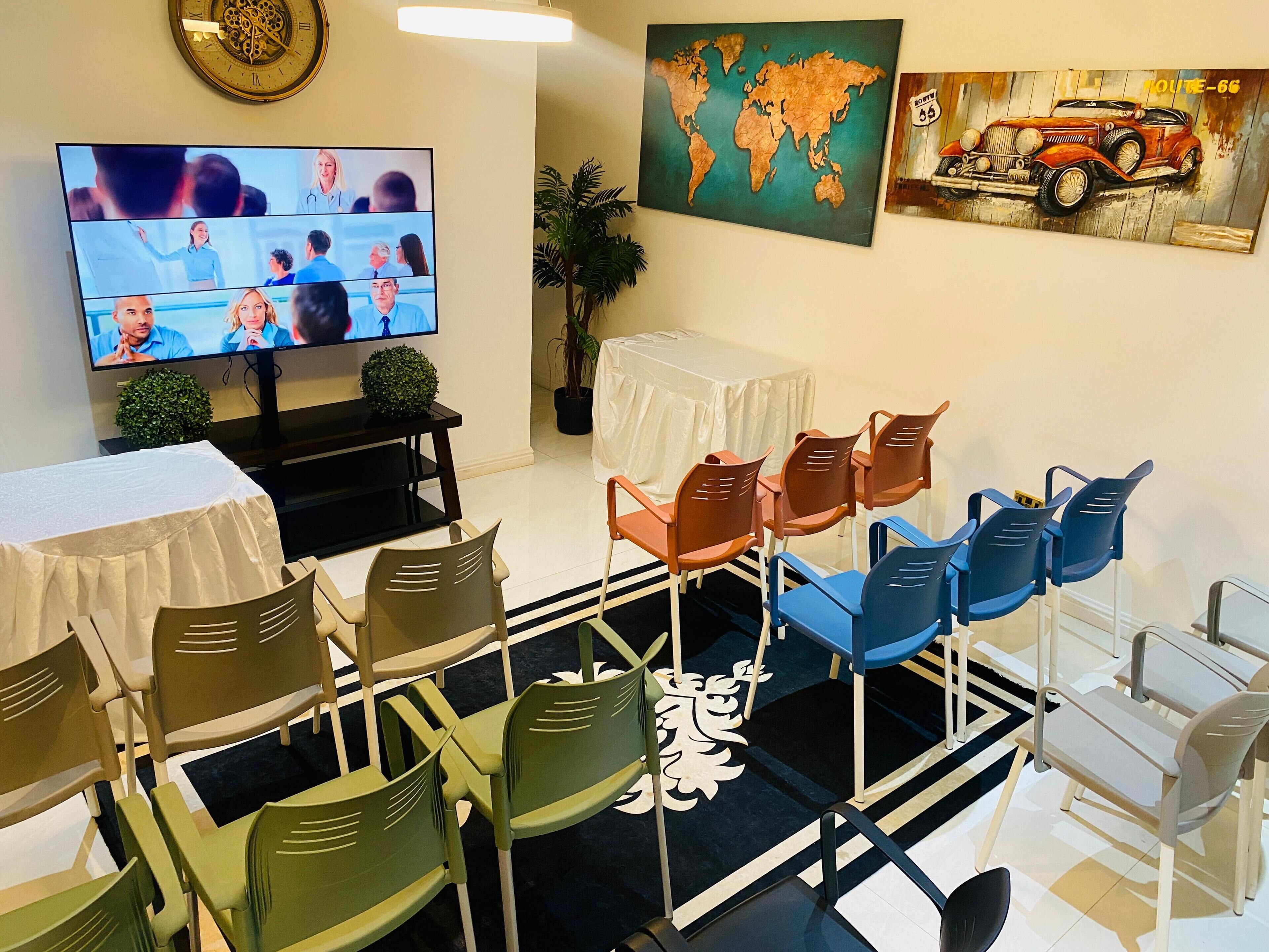 A conference room with a large flat-screen TV displaying a meeting, surrounded by chairs and a small table.