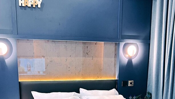 A modern hotel room with a large bed, white bedding, a headboard with integrated lighting, and a 'HAPPY' sign above the headboard. - Wallgate Apartments (Abuja)