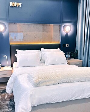 A modern hotel room with a large bed, white bedding, a headboard with integrated lighting, and a 'HAPPY' sign above the headboard. - Wallgate Apartments (Abuja)