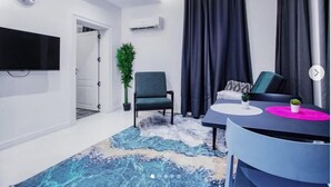 A modern living room with a flat-screen TV, a potted plant, a grey armchair, a round coffee table with a pink placemat, and a patterned rug. - Wallgate Apartments (Abuja)