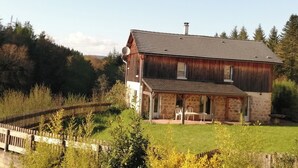 Ein rustikales, zweistöckiges Haus mit Steinunterbau und einer Metallbedachung, umgeben von einem Holzzaun und üppiger Vegetation.