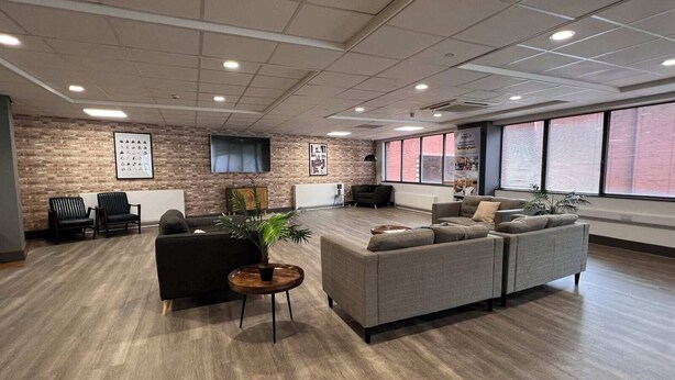 A spacious lobby with a modern design, featuring a large grey sectional sofa, a small round table with a plant, and a brick wall with a mounted TV.