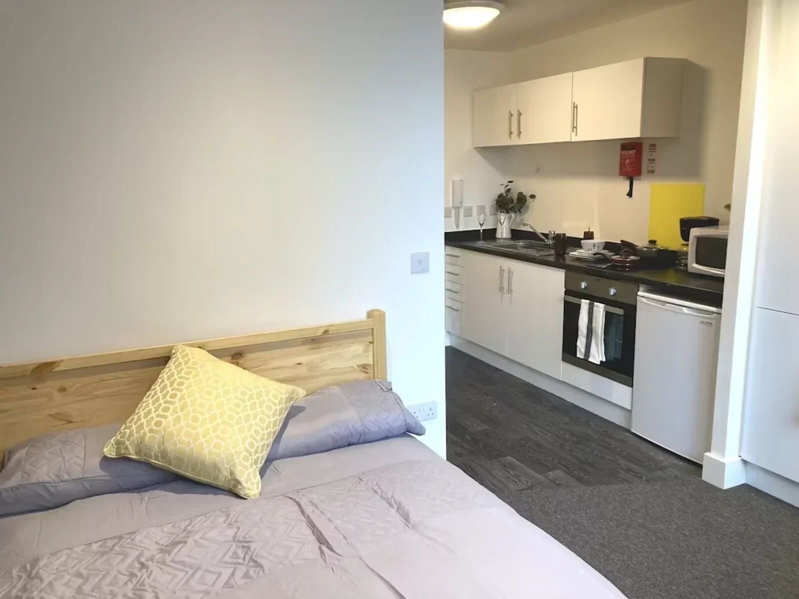 Un dormitorio con cabecera de madera y cama con ropa de cama estampada. Junto a ella, una kitchenette con armarios blancos, un microondas y un lavamanos.