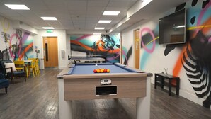 A pool table with balls in a room with wooden flooring, a dining area with yellow chairs, and a mural on the wall.