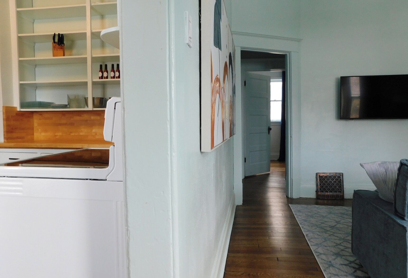 A modern kitchen with white cabinetry, a built-in oven, and a wooden countertop. There is a hallway leading to a living area with a gray sofa and a flat-screen TV mounted on the wall.