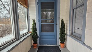 A blue front door with a glass panel, flanked by two potted evergreen shrubs, leading to a black textured mat.