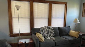 Una sala de estar con un sofá gris, una mesita de madera, una lámpara de pie y una ventana con persianas.