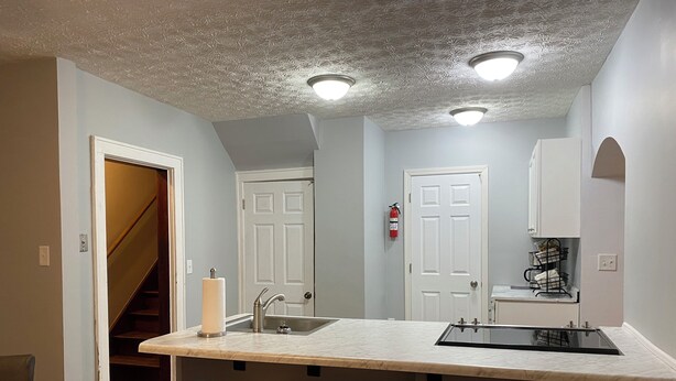 Una cocina con encimera de mármol, dos taburetes de madera, un lavamanos de acero inoxidable y una puerta blanca con extintor.