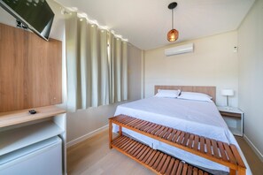 A bedroom with a bed, a TV mounted on the wall, a wooden bench, a bedside table, and a lamp. - Suíte Ala pôr do sol Residencial Solavir (Bombinhas)