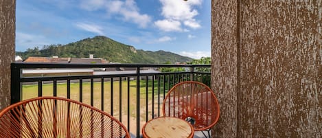Un balcón con dos sillas de mimbre y una mesita pequeña, con vistas a un paisaje verde y una montaña.