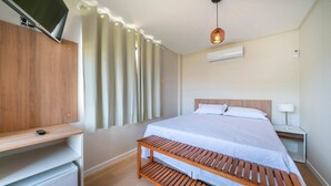 Quarto com cama, televisão na parede, um banquinho de madeira, mesa de cabeceira e abajur.