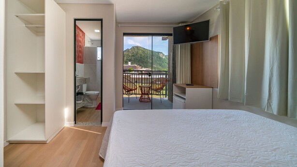 Quarto de hotel com uma cama, televisão, varanda com cadeiras e vista para uma montanha.