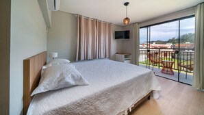 Quarto com cama grande, televisão na parede e varanda com mobiliário para exterior.