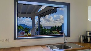 A kitchen with a large window overlooking a scenic landscape, a sink, and a coffee maker. - Villa Paruline with swimming pool tropical garden and sea view (Grand-Bourg)