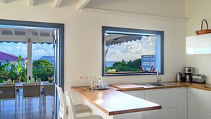 A modern kitchen with a large window, a dining area with chairs, and a view of the outdoors. - Villa Paruline with swimming pool tropical garden and sea view (Grand-Bourg)