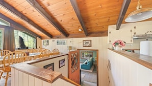 Interior rústico de una cabaña con vigas de madera, comedor con sillas, cocina con armarios y una sala acogedora con sofá.